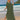 A woman wearing an elegant green V-neck caftan stands on a sandy beach with a clear sky and azure water in the background.