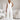 Woman modeling an elegant white sleeveless V-neck belted formal jumpsuit, showcasing the shoulder, neck, waist, and thigh areas, captured with flash photography.