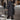 Women's long black insulated winter down coat, featuring a stylish urban design. The coat extends to the knees and includes full-length sleeves, designed for warmth in snowy conditions. It has a waist-cinching fit, resembling a trench coat style, suitable for fashionable outerwear.