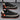 Men's urban black sneakers with contrast orange detailing, featuring a cushioned heel and ribbed sole, displayed against a white background.
