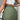 Olive Green Women's Utility Mini Cargo Skirt with a Belt and Pockets, modeled by a person wearing a white top. The skirt features a stylish utility design with practical pockets and a comfortable waistband.