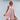 A smiling woman wearing a pink linen tiered mini dress with a sleeveless design and a V-neckline, perfect for summer fashion. The dress, in a rose color and size XL, features a flattering waist and falls gently above the knee.