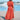 A woman wearing a vibrant red maxi beach dress stands outdoors against a backdrop of clear blue sky and water. The lightweight summer dress features a side slit, showcasing a flowing, elegant design with subtle orange undertones. The dress has a fitted waist and sleeveless neckline, perfect for a warm day.