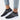 A pair of women's fashion slip-on sneakers featuring a black mesh design adorned with rhinestones, displayed on a person's feet. The sneakers are styled with casual grey sportswear and denim, highlighting their versatility and trendy appeal.