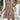 Woman wearing short summer dress with geometric print, keyhole neckline, and tassels, standing with hand on hip, showcasing shoulder and hairstyle with plants in the background.