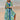 A woman wearing a tropical print green beach kaftan, standing near water. The lightweight V-neck summer dress features short sleeves and a vibrant pattern, perfect for a sunny day.