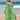 A woman wearing a tropical print green beach kaftan stands near the water, smiling under a clear blue sky, showcasing the lightweight V-neck summer dress with loose sleeves and a relaxed waist.