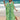 A woman wearing a Tropical Print Green Beach Kaftan, a lightweight V-neck summer dress, standing on a beach near the water, with her arms raised and a happy expression.