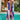 Woman wearing a vibrant print kaftan, standing near water and surrounded by tropical plants. The dress features purple and aqua colors, designed for luxury resort and tropical beach wear, showcasing stylish shoulder and sleeve details.