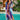 A woman wearing a vibrant print kaftan dress with tropical and luxurious resort styling, posing against a lush green background with houseplants and water elements.