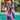 Woman wearing a vibrant print kaftan dress with purple and tropical patterns, standing near a window with houseplants in the background. The dress features a loose, flowing design with a focus on leisure and resort fashion.