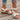White floral wedge sandals featuring a beaded strap and elegant cutout design, shown on a model's foot and leg, highlighting the shoe's beige color and stylish appearance.