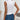 A woman standing and wearing a white stretch cotton sleeveless top with ruched detailing, paired with blue jeans. The focus is on the top's fit around the neck and shoulders, highlighting its summer-appropriate style. The background is a light curtain, adding a soft, casual ambiance.
