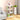 White X-shaped corner bookshelf with a modern minimalist design, featuring wooden shelves and a variety of items such as houseplants, a flowerpot, and toys displayed on it. The bookshelf is situated on wooden flooring.