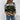 A woman wearing a green, black, and beige camouflage knit sweater styled in a street fashion manner, paired with jeans.