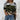Women's street style knit sweater in a camouflage pattern with shades of green, black, and beige, featuring long sleeves and a relaxed fit, styled with jeans.