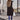 A woman wearing a long black puffer vest styled as an urban chic winter jacket, featuring a sleek design with a waist-length fit. She is standing, showcasing the vest's shoulder and sleeve details.