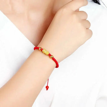 A hand wearing a 24K gold red string bracelet with 3D Pixiu beads, showcasing the 999 gold stamp on the wrist.