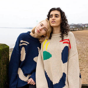 
A woman wearing an oversized knit sweater with an abstract face design, characterized by a cozy and artistic fashion style. She is outdoors, showcasing the sweater's long sleeves and relaxed fit, with a cheerful expression and stylish hairstyle.