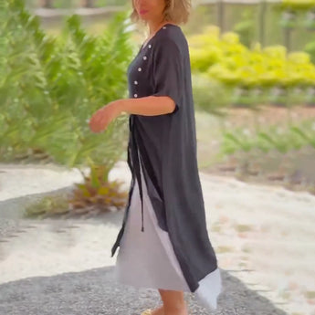 Asymmetrical blue V-neck maxi dress with side buttons, showcasing a sleeveless design and cinched waist, worn by a person with their hand gesturing at their side.