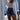 Women's summer fashion featuring a beige knit crop top paired with white shorts. The outfit is styled with a hat and set against an azure background, emphasizing chic beachwear. The ensemble highlights the shoulders and waist, reflecting a casual and trendy look.