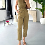 Beige sleeveless belted jumpsuit displayed on a mannequin with a focus on casual chic summer fashion, featuring a defined waist and flowing design, set against a backdrop with houseplants and a window.