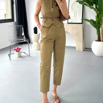 Beige sleeveless belted jumpsuit displayed on a mannequin with a focus on casual chic summer fashion, featuring a defined waist and flowing design, set against a backdrop with houseplants and a window.