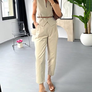 Beige sleeveless belted jumpsuit displayed on a hanger, featuring a chic summer design with a belt at the waist, set against a neutral background with a potted plant nearby.