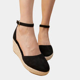 
A pair of black wedge espadrilles with a closed toe and ankle strap, crafted from suede material, displayed in a fashion-forward setting.