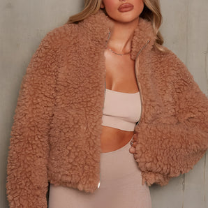 
A beige teddy coat for women in size 3XL, featuring a fluffy texture with a zip-up front. The trendy jacket has long sleeves and a high neck, designed for warmth and style, displayed in a camel color.