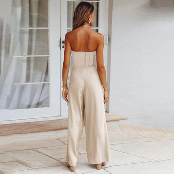 A beige sleeveless jumpsuit featuring a tie-waist design, suitable for elegant casual summer wear. The jumpsuit highlights areas such as the neckline, waist, and thighs, and is showcased against a wooden floor background.