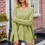 
A woman wearing a green oversized batwing poncho sweater stands indoors. She has an elegant hairstyle and is accessorized with sunglasses. The setting includes a window and houseplants, giving a cozy and stylish fall fashion vibe.