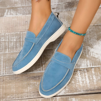 Beige women's suede slip-on loafers worn on feet, shown in a casual setting with a blue background.