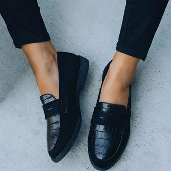 Women wearing black crocodile loafers, luxury designer slip-on shoes, paired with a stylish grey dress.