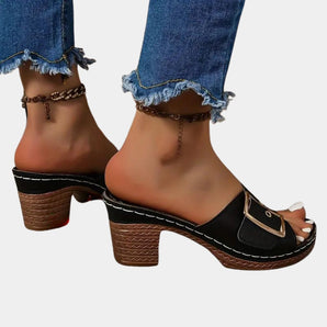 Black heeled dressy sandals featuring a buckle strap and an elegant ankle chain detail, worn on a woman's foot, highlighting the shoe's stylish design and high heel.