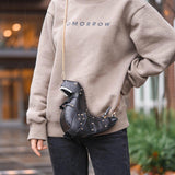 Black leather horse-shaped crossbody bag with a gold chain strap displayed against a backdrop of fashionable clothing items and accessories.