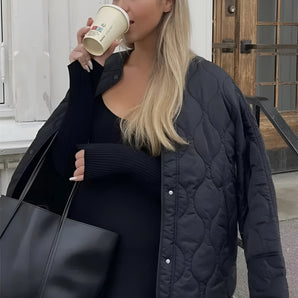 Women's black oversized quilted jacket with long sleeves, featuring a lightweight urban puffer design; model standing with a casual gesture showcasing the jacket's fashionable outerwear style.