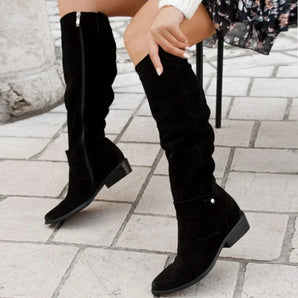 Black suede knee-high flat heel winter boots featuring a side buckle, shown on a model’s leg.