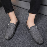 Men's black textured leather slip-on loafers with zipper, shown on a person wearing blue denim jeans, standing on a grey floor surface.