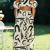 
Fashion model wearing a black and white abstract two-piece dress consisting of an off-shoulder top and palazzo pants, styled with sleek hair and white shoes, standing on grass. The dress emphasizes the waist and thigh areas.