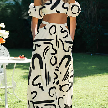 
Fashion model wearing a black and white abstract two-piece dress consisting of an off-shoulder top and palazzo pants, styled with sleek hair and white shoes, standing on grass. The dress emphasizes the waist and thigh areas.