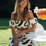 Off-shoulder black and white abstract two-piece dress featuring a smiling woman posing outdoors, highlighting the stylish fashion ensemble with palazzo pants amidst a natural grassy backdrop.