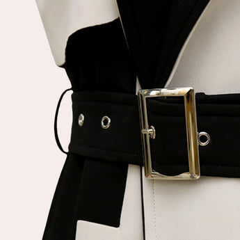 Black and white belted trench coat with a monochrome chic design, featuring a collar, belt with buckle, and long sleeves, worn by a person who is also wearing glasses and holding a bag, seated on a chair.