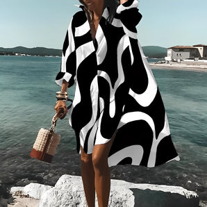 
A woman wearing a black and white geometric maxi dress styled as a bold graphic beach cover-up, standing near a lake under a clear sky. The dress features a long sleeve design and covers the thighs, suitable for a beach setting. The woman appears happy and confident, embodying a sense of fashion and style.
