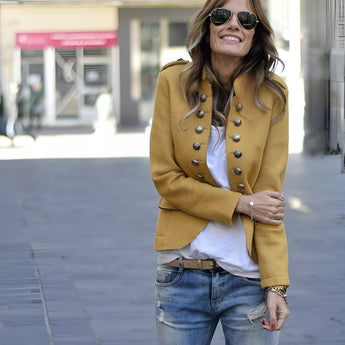 A woman wearing a black double-breasted military jacket with silver buttons, paired with jeans and sunglasses, smiling.