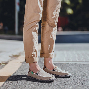 A pair of black woven loafers designed as breathable and lightweight slip-on shoes, showcased on a wooden floor with a person wearing them alongside a pair of jeans.