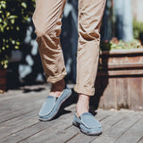 Black woven loafers shown worn by a person standing on a road surface, paired with grey denim jeans.
