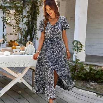 Blue floral V-neck wrap maxi dress with short sleeves, worn by a model standing near plants, showcasing the dress's elegant design suitable for summer and garden parties.