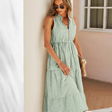 Blue and green striped sleeveless midi dress with a V-neck and button front, worn by a woman standing near a window, accessorized with sunglasses.