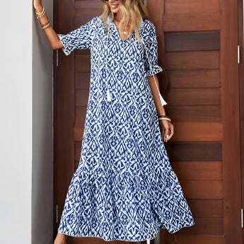 A woman wearing a blue and white V-neck boho maxi dress with short sleeves, standing outdoors on a sunny day. She is accessorized with sunglasses and sandals, embodying a lightweight summer beachwear style.